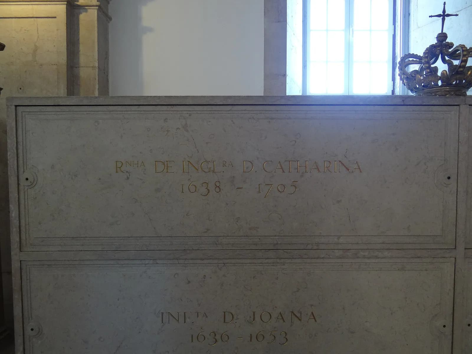 Two stacked tombs of pale grey stone with gilded lettering in a white room. The upper one reads Rnha de Inglra D. Catharina, 1638-1705; the lower one reads Infta D. Joana, 1636-1653. A gilded crown with a cross sits on top.