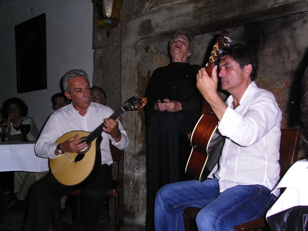 An elderly woman in a long black dress stands singing with her eyes half closed against a rough stone wall, between two seated guitarists in white shirts. The man on the left plays a pear-shaped Portuguese guitar, the man on the right a classical guitar. Diners sit at a table behind them.