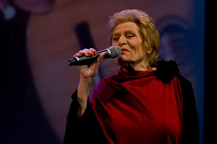 An elderly woman with short fair hair and red nail varnish sings into a hand-held microphone, wearing a deep red shawl over a black dress, against a dark stage backdrop.