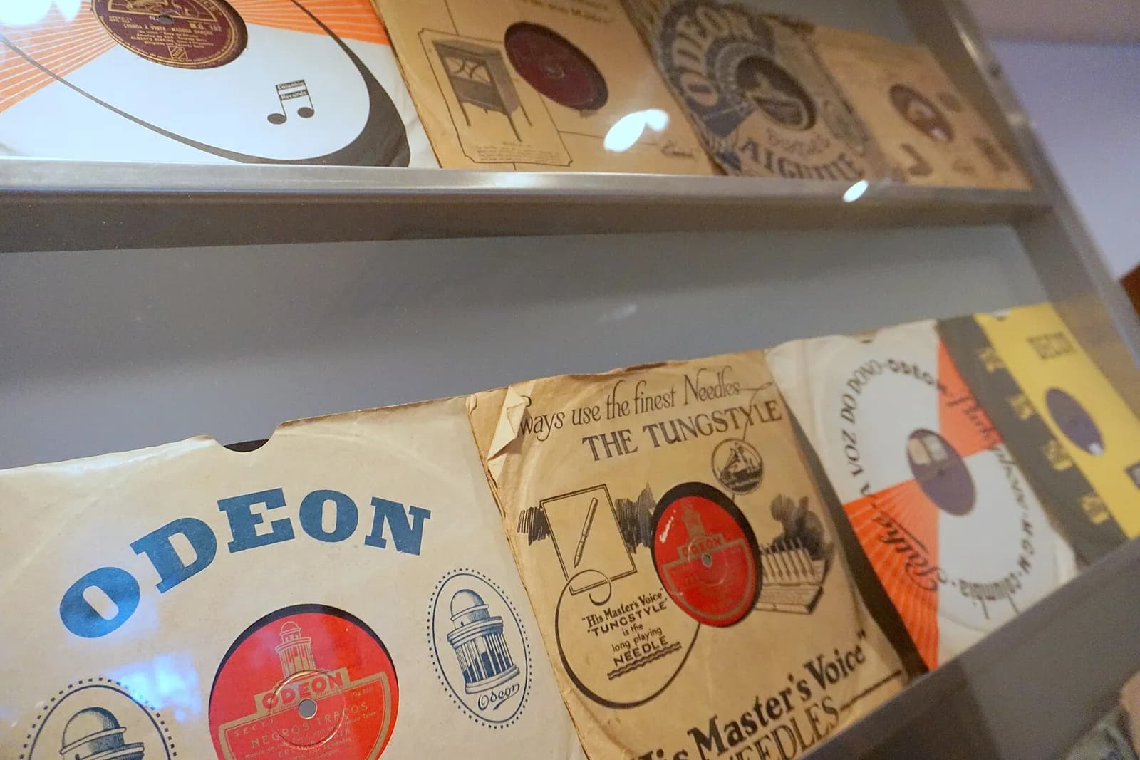 Old 78 rpm records in worn paper sleeves printed with Odeon, His Master's Voice, Columbia and Decca logos, displayed on two shelves behind glass.