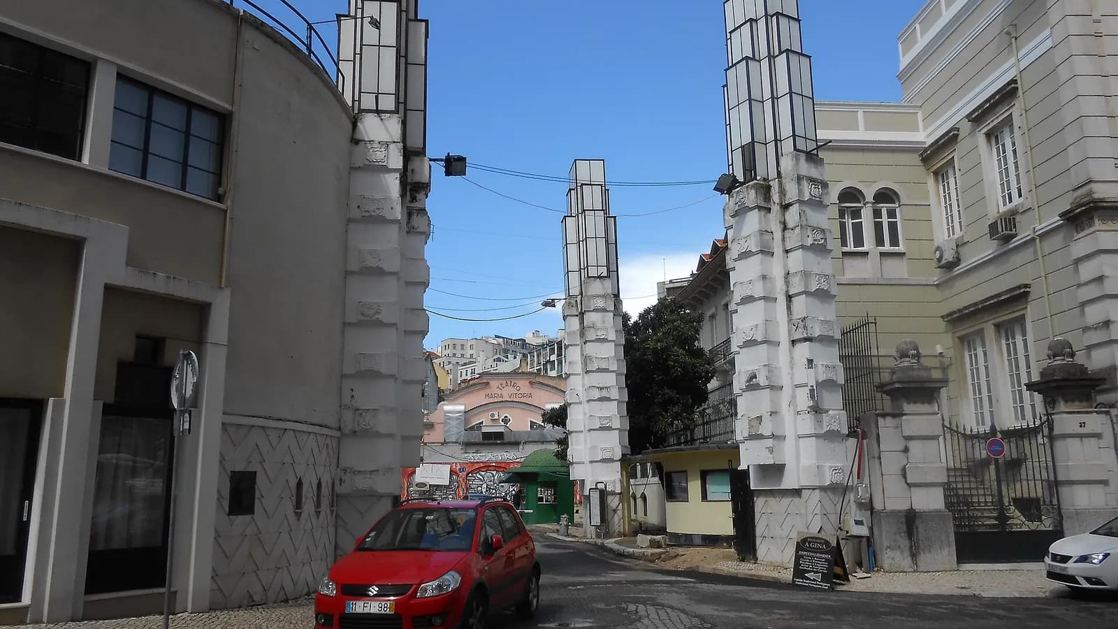 A street passes between tall white stone pillars topped with glass lanterns, leading towards a pink theatre front marked Teatro Maria Vitória. A red car is parked in the foreground.