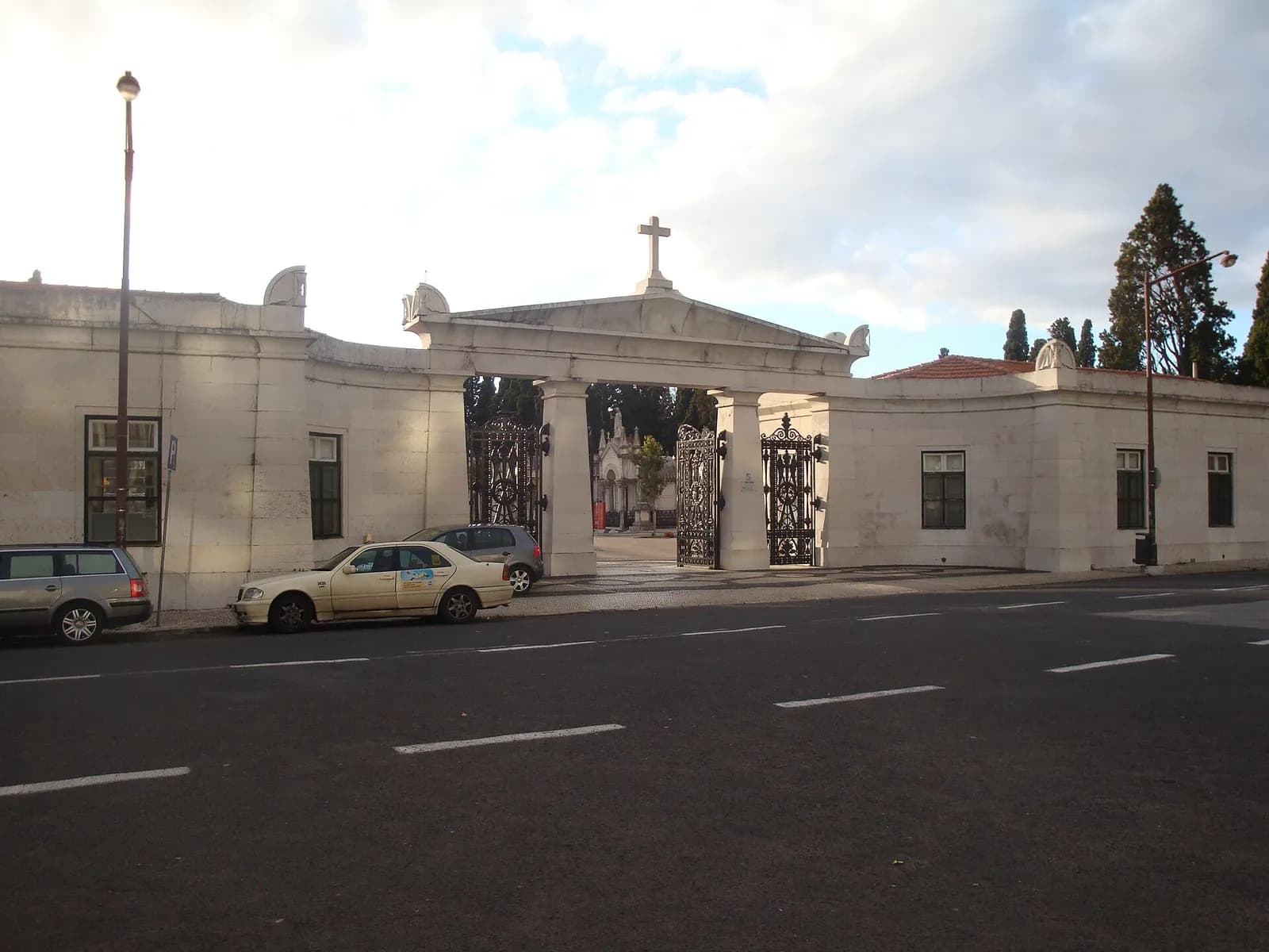 A long, low white stone wall with a gabled gateway topped by a cross and open wrought-iron gates, with tombs and cypress trees visible behind it. A taxi and two cars are parked on the street outside.