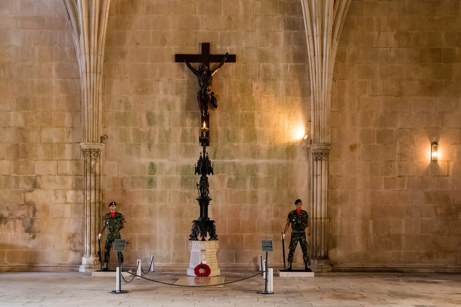 Two soldiers in camouflage uniform and berets stand guard on either side of a dark bronze crucifix on a stone wall in a vaulted hall. Below the cross a tall ornate black lamp holds a small flame above a stone plinth with a red wreath.