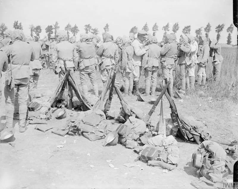 Soldiers in hooded gas masks stand in a crowd on a dusty field, their rifles stacked in small pyramids and packs and helmets scattered on the ground in front of them. A line of tall poplars runs along the horizon.