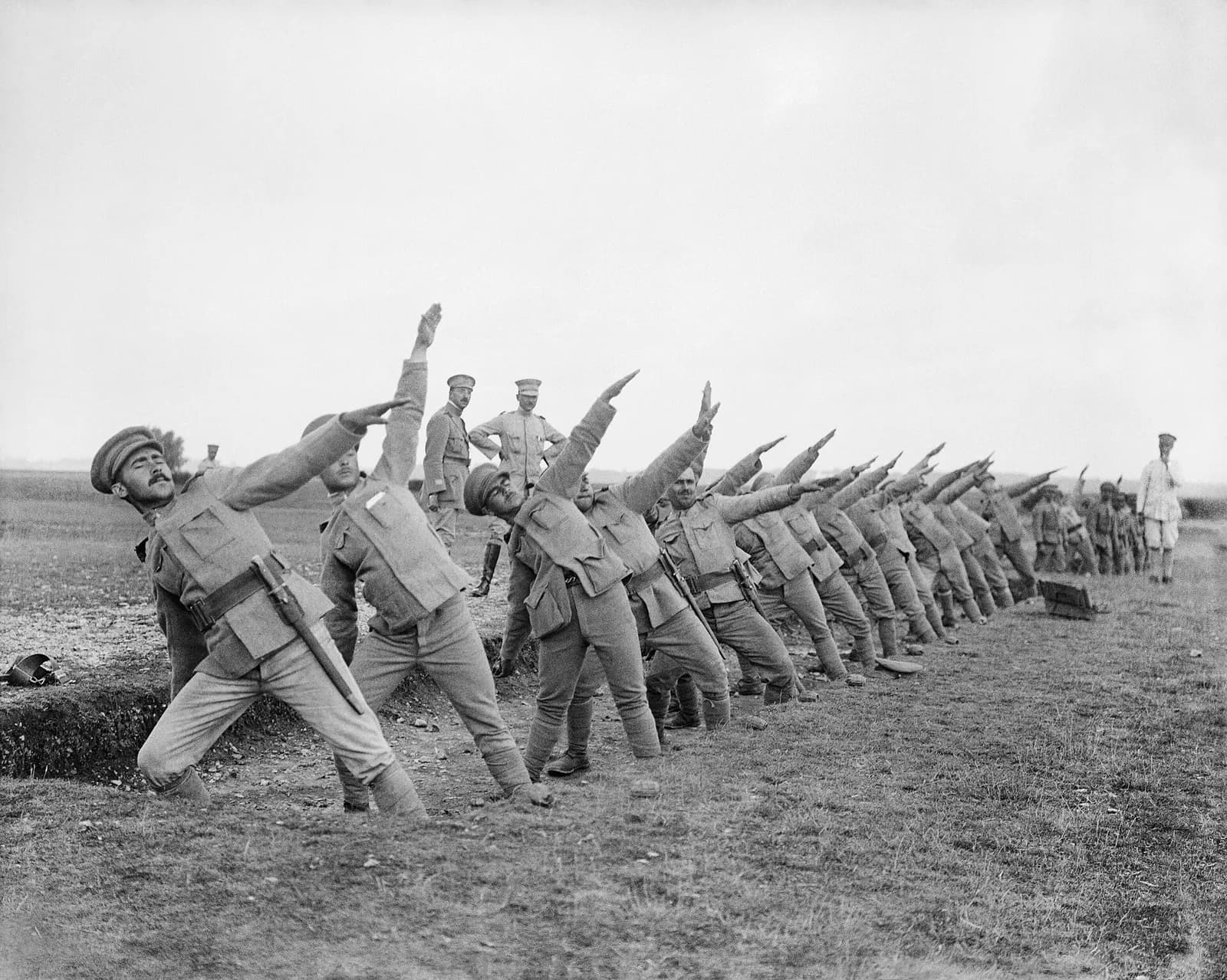 A long line of soldiers in pale uniforms, caps and puttees leans back on a bare field, each with one arm flung high as if about to throw. Two officers, one with his hands on his hips, watch from behind the line.