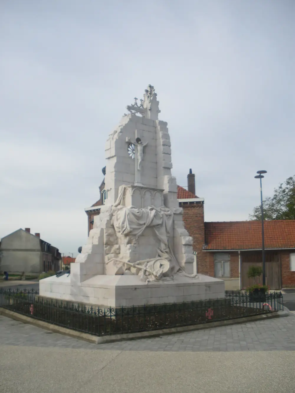 A white stone monument shaped like the ruined wall of a Gothic church, with a rose window and a crucifix, stands behind an iron railing on a village square. Draped cloth, a fallen helmet and weapons are carved at its base, and red-roofed brick houses stand behind it.