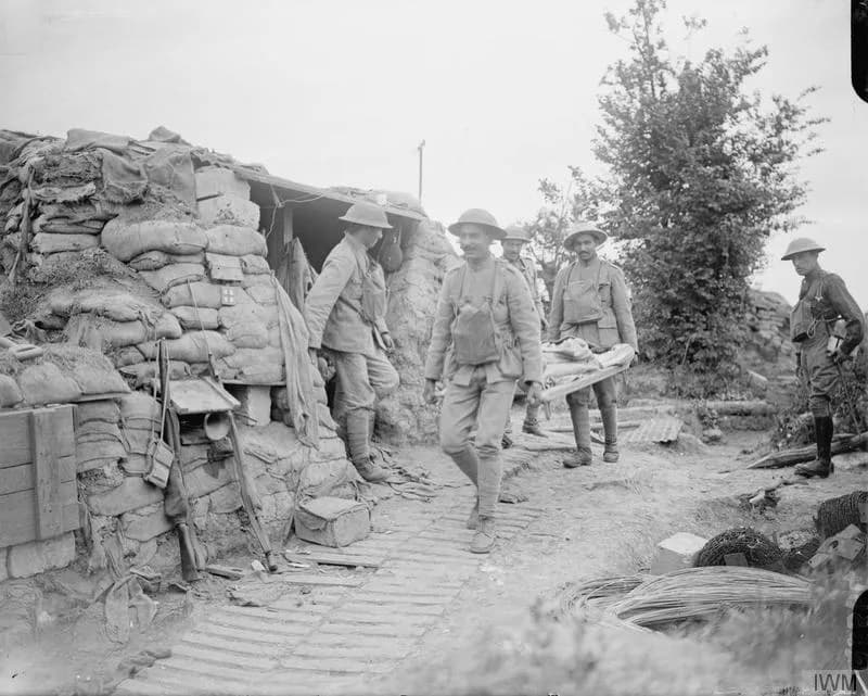 Soldiers in steel helmets carry a man on a stretcher along a duckboard path past a dugout walled with stacked sandbags. Coils of wire lie beside the path and a small tree grows behind the sandbag wall.