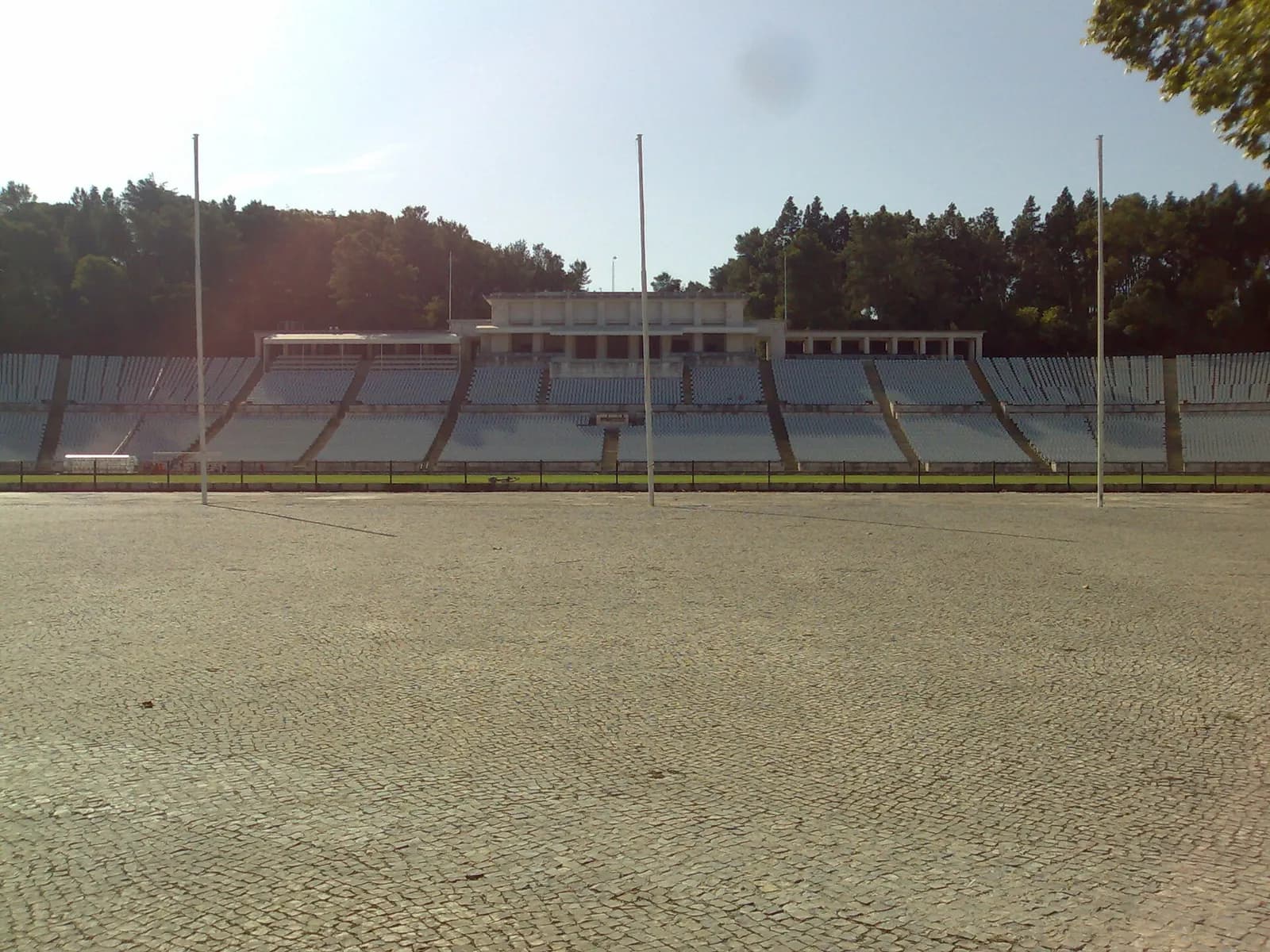 Empty white-seated stands of a large open-air stadium with a colonnaded central tribune, three tall white flagpoles, woods behind and a wide cobbled square in the foreground.
