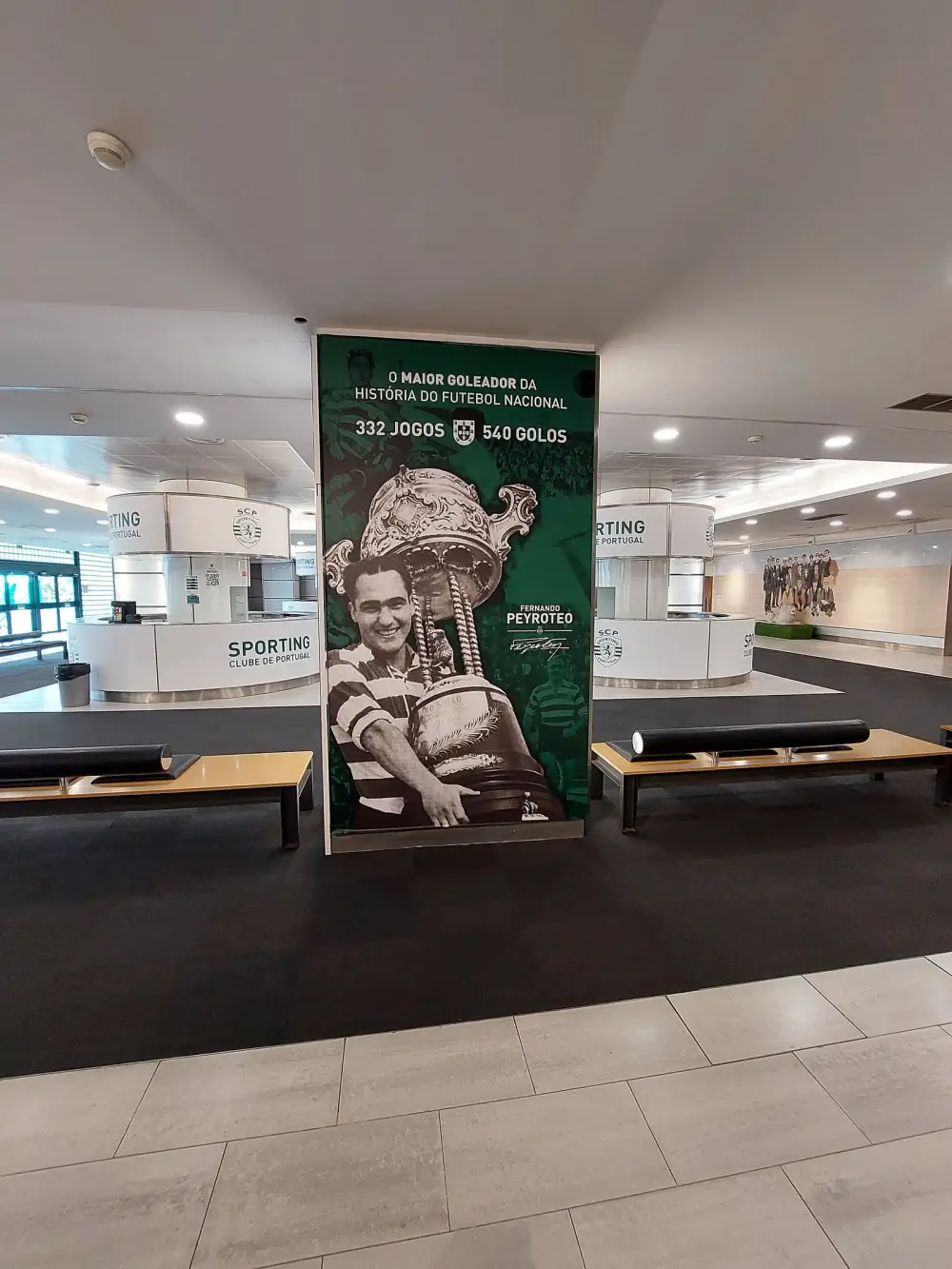 A tall green panel in a stadium lobby shows a black-and-white photograph of a smiling footballer in a hooped shirt holding a huge ornate trophy, under the words O maior goleador da história do futebol nacional, 332 jogos, 540 golos.