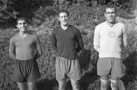 Three footballers stand side by side on a grassy bank: a slim young man in a dark shirt and shorts, a stockier man in a dark shirt, and a goalkeeper in a light jersey with a cross badge and knee pads.