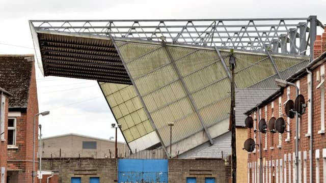 The back of a large football stand with a sloping corrugated roof and steel frame rises above a row of red-brick terraced houses with satellite dishes.