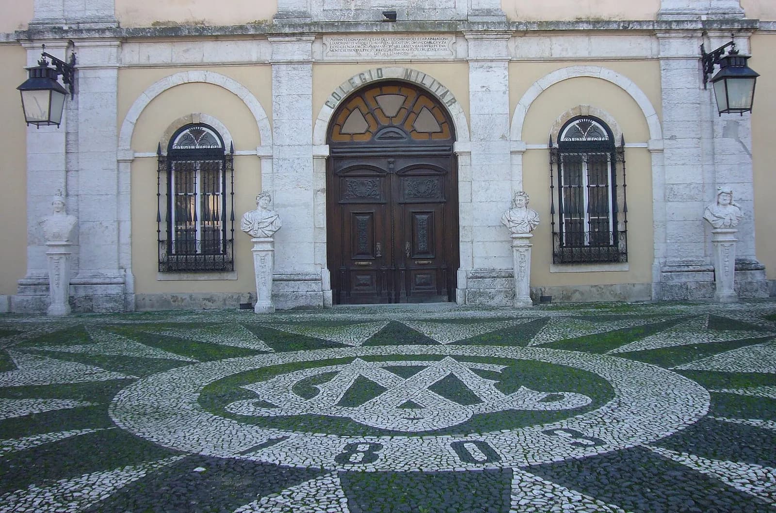 An arched wooden double door under the words Colégio Militar, flanked by barred windows and four marble busts on pedestals. A long carved inscription runs above. In front, a black-and-white cobbled pavement shows a large monogram and numerals.