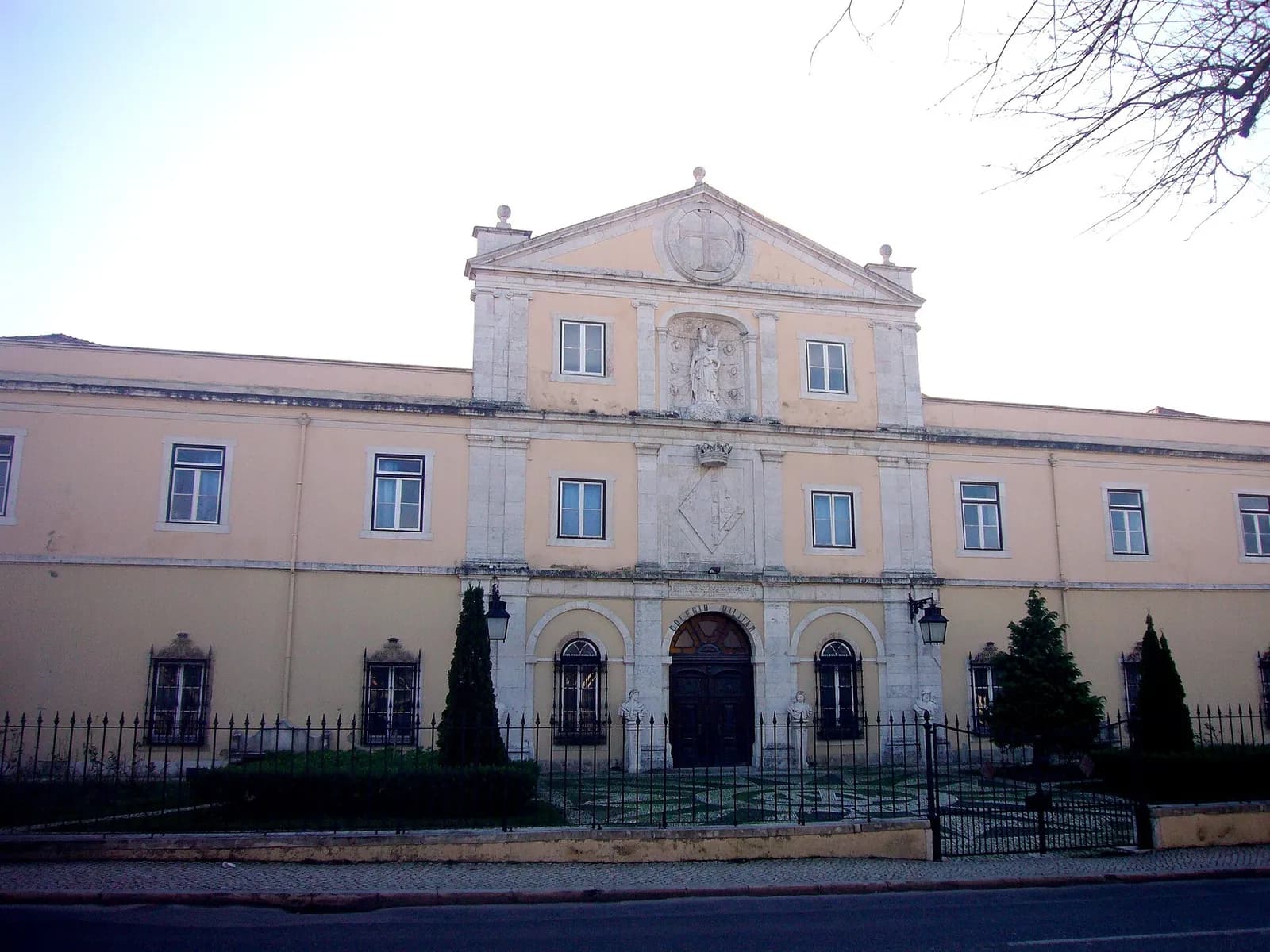 A long, two-storey ochre building behind iron railings. The centre rises into a pediment with a carved cross, a statue in a niche and a coat of arms over an arched doorway.