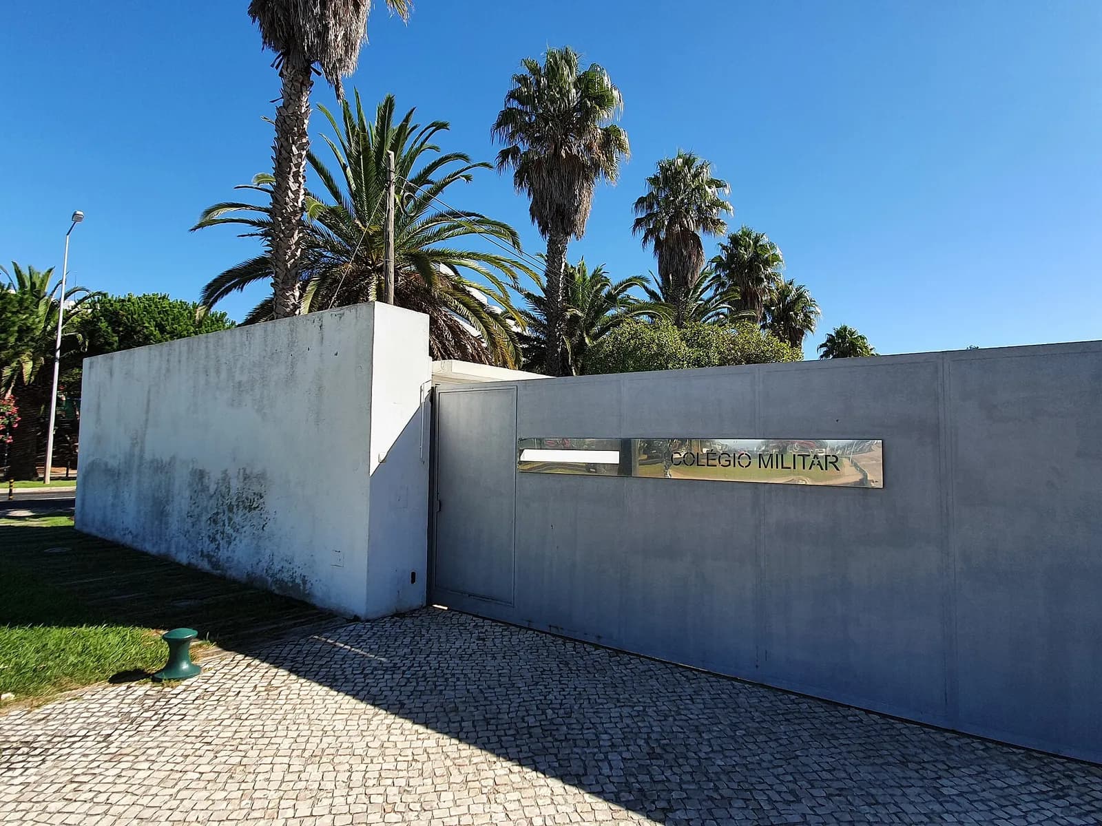 A plain grey concrete wall and gate with a steel plaque reading Colégio Militar, under tall palm trees and a clear blue sky.