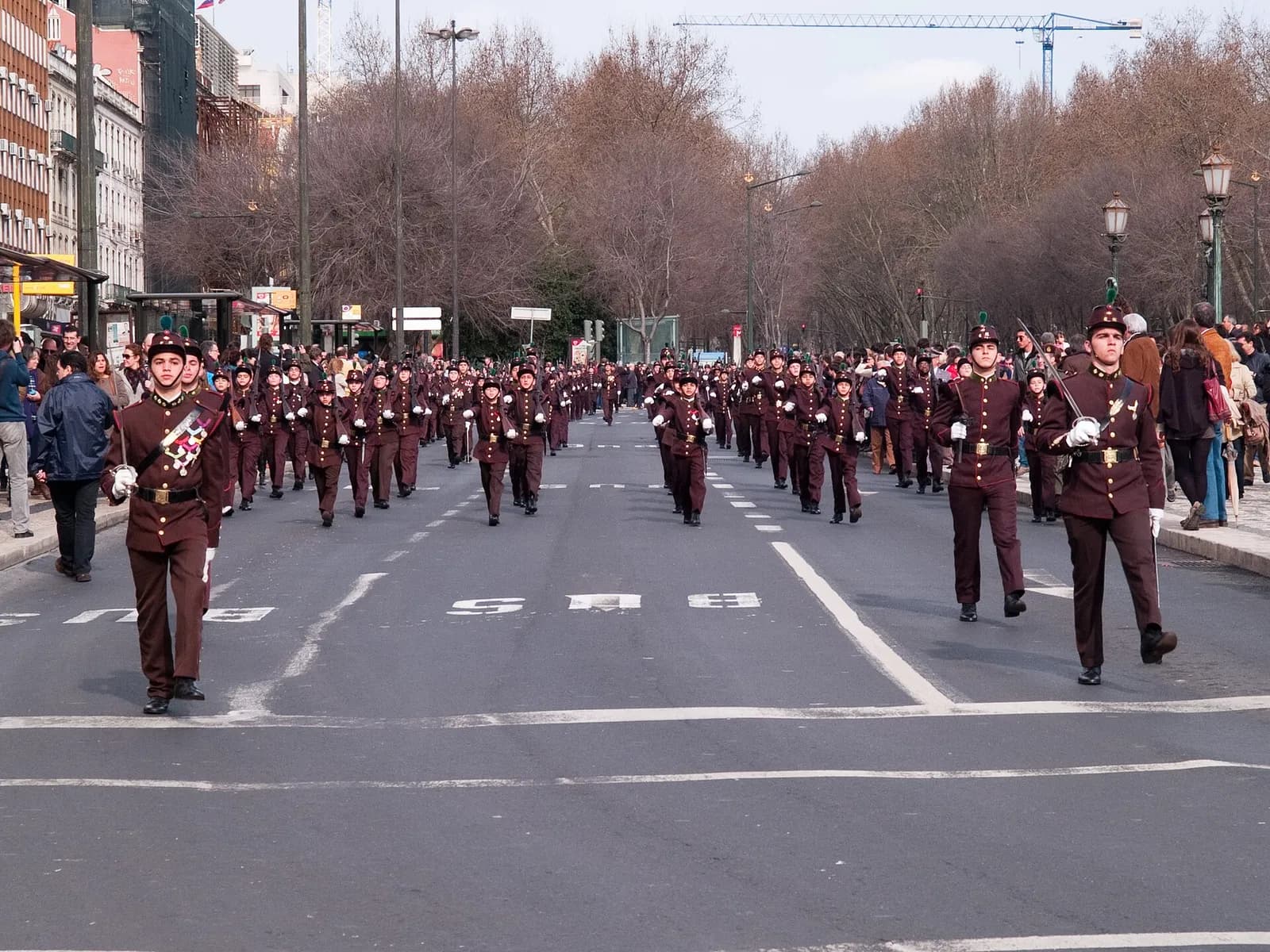 Rows of young cadets in brown uniforms, white gloves and plumed shakos march down a wide tree-lined avenue, led by cadet officers with drawn swords, while spectators watch from the pavements.