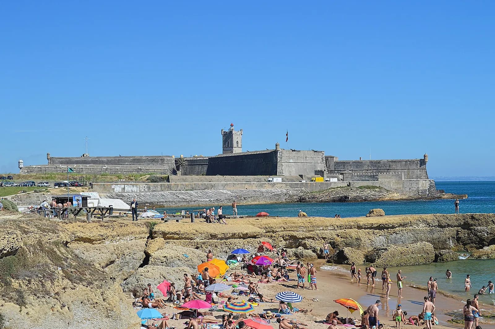 A low grey stone fortress with a square tower and battlements stands on a rocky point above the sea, with a crowded sandy beach and bathers in the foreground.