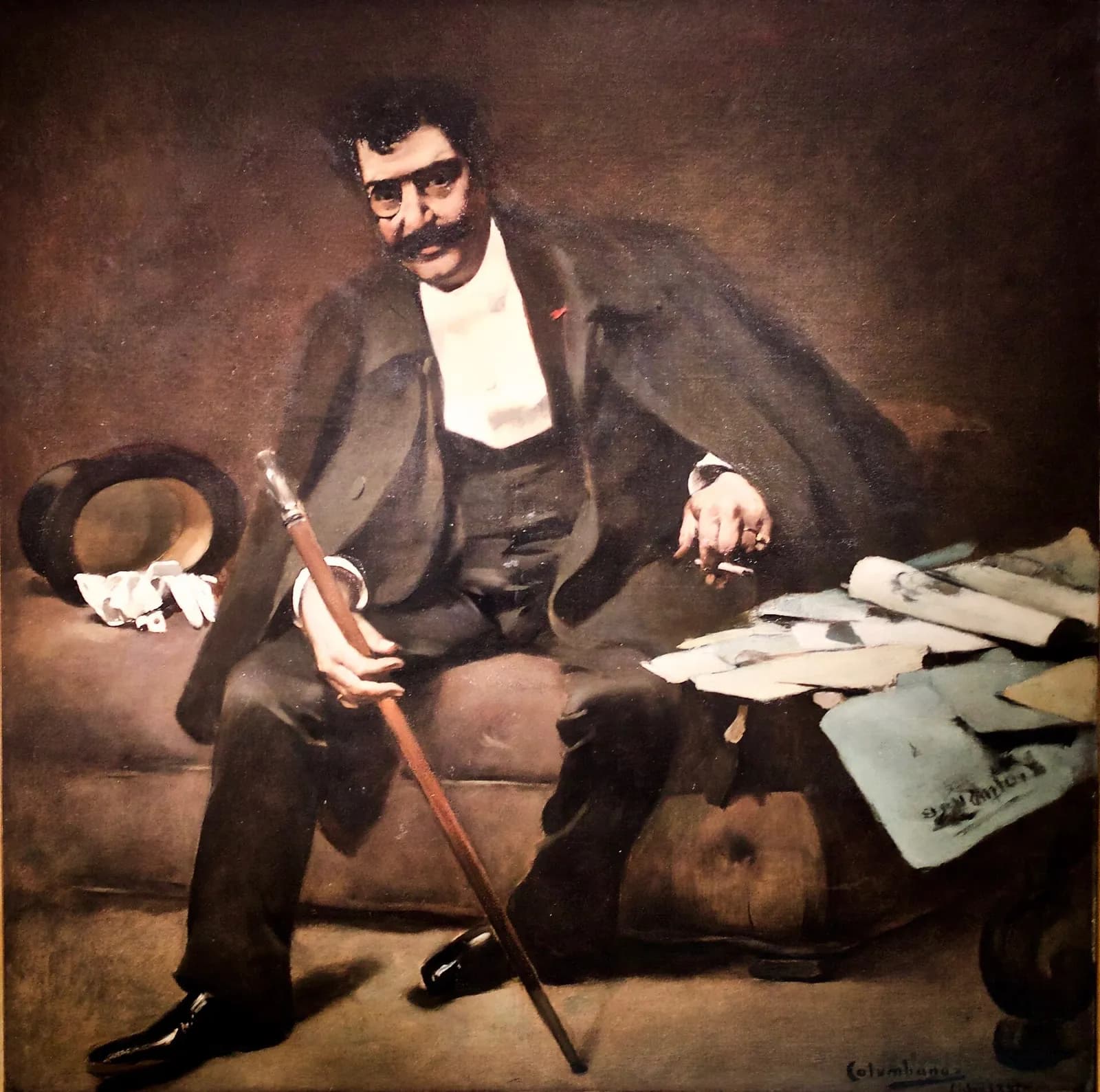 A full-length painting of a man with dark curly hair, a pince-nez and a thick black moustache, sitting on a brown sofa in a dark coat and white shirt. He holds a long walking cane in one hand and a cigarette in the other. A top hat and white gloves lie beside him, and newspapers and drawings are piled at the right.