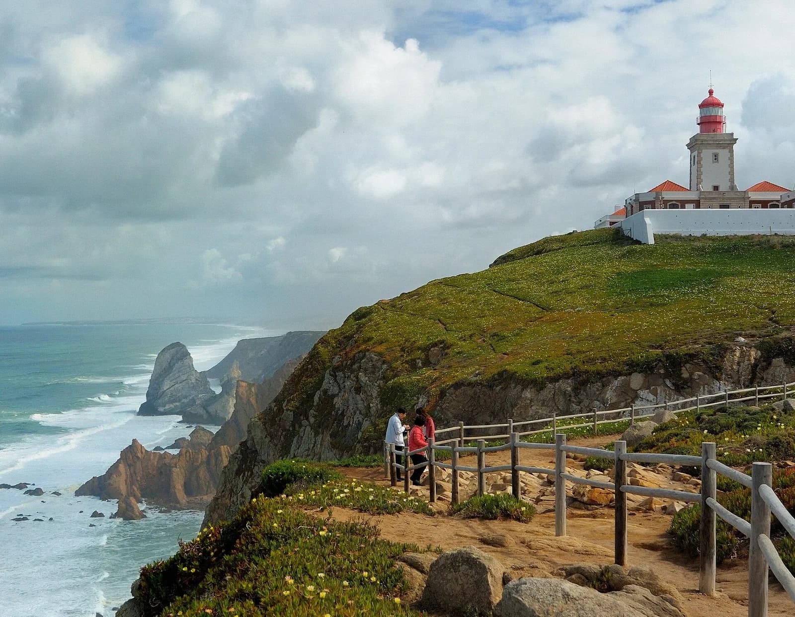 Green clifftops above a rough Atlantic under heavy cloud, with jagged rock stacks in the white surf below and a stone lighthouse with a red lantern above white walls on the headland. A few visitors stand at a wooden fence on the clifftop path.
