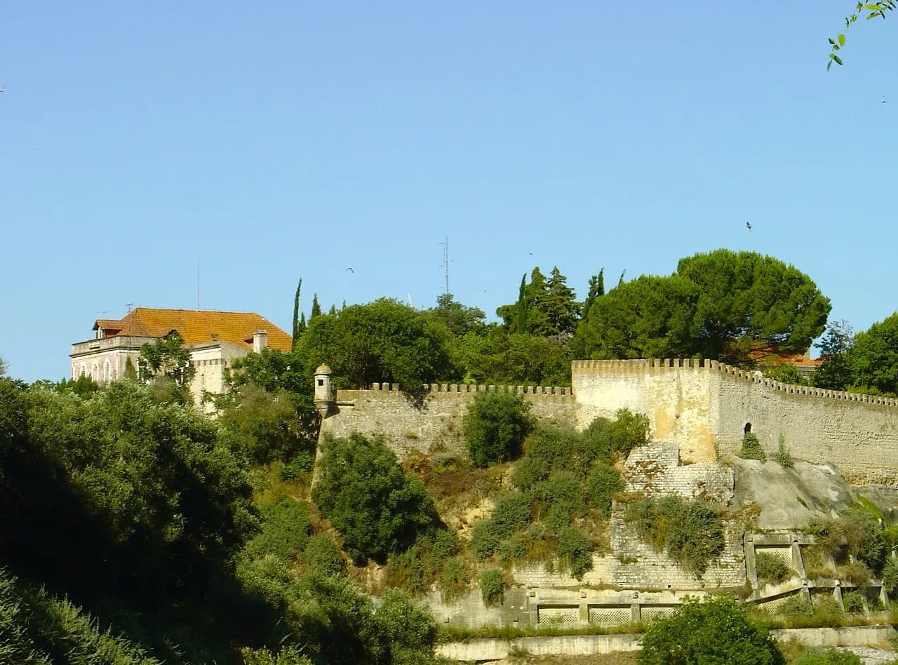 A long crenellated stone wall with a small round sentry box runs along the top of a steep, overgrown slope, with pine trees and a large tiled house behind it under a clear sky.