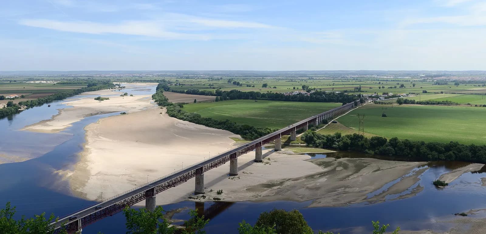 A wide view from a height over a broad river with sandbanks, crossed by a long iron bridge on stone piers, and flat green fields stretching to the horizon.