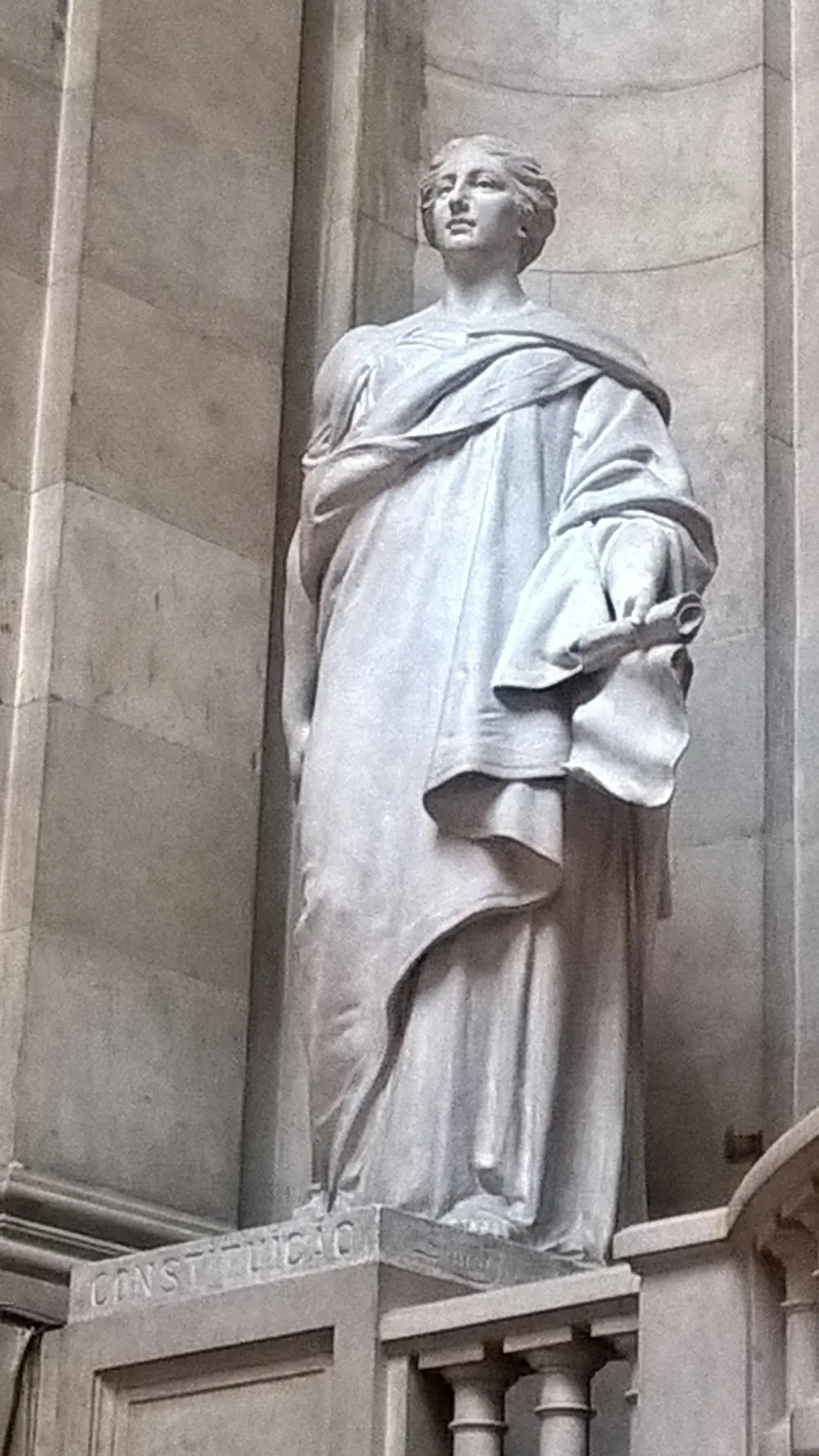 A white plaster statue of a woman in classical robes holding a scroll, standing in a stone niche on a pedestal inscribed CONSTITUIÇÃO.