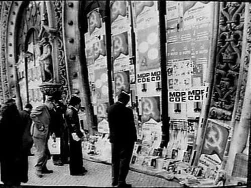 Black-and-white photograph of people in coats standing in front of a carved stone façade covered with election posters, among them hammer-and-sickle posters and posters for the MDP/CDE.