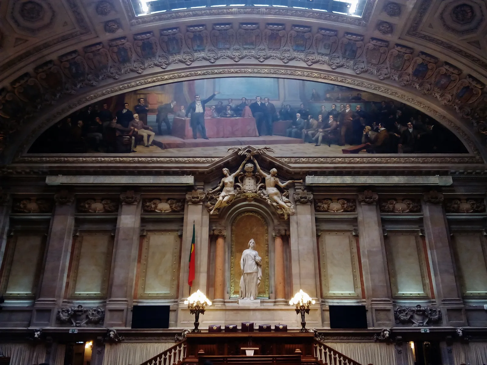 The end wall of an ornate parliamentary chamber: a large arched painting of a nineteenth-century assembly above a marble wall with a white statue in a gilded niche, a Portuguese flag, two lamps and the wooden presidential bench below.