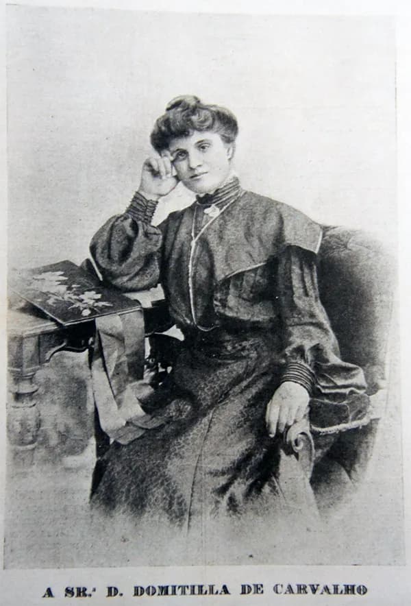 Printed portrait of a young woman with her hair piled up, in a dark high-collared dress with puffed sleeves. She sits beside a small table with her elbow resting on it and her head against her hand. The caption below reads “A Sr.ª D. Domitilla de Carvalho”.