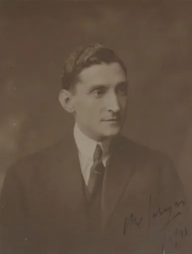 Sepia studio portrait of a slim man with dark, wavy hair, in a dark suit and tie, looking to his left. A signature and the year are written across the lower right corner.