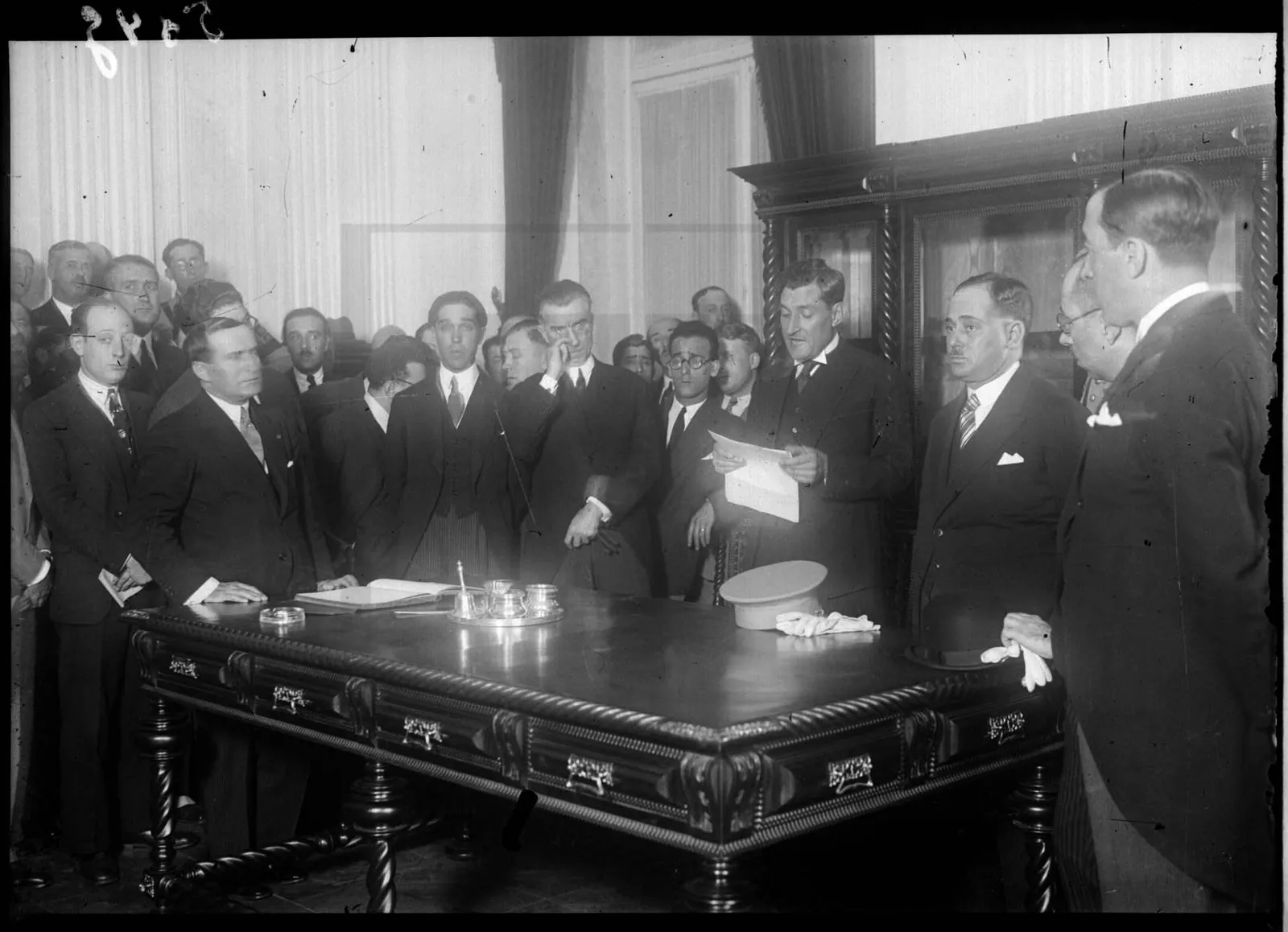 Black-and-white photograph of a crowded room. A man in a dark suit reads aloud from a sheet of paper behind a large carved wooden table, on which lie an open book, an inkstand, a military cap and white gloves. Men in suits stand pressed around him.