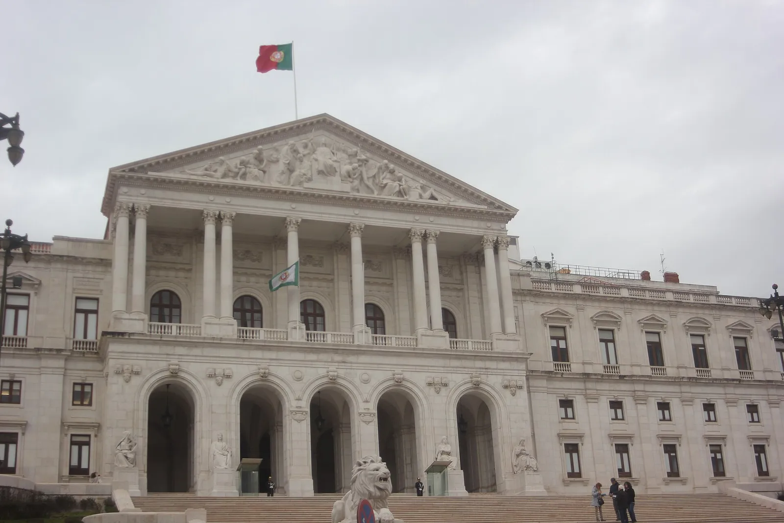 The white neoclassical front of the Palácio de São Bento under a grey sky, with a columned portico, a sculpted pediment, five arches at ground level and a wide flight of steps with a stone lion at its foot. The Portuguese flag flies from the roof.