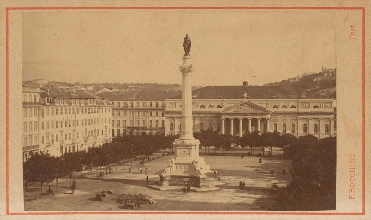 A sepia photograph of a large paved square lined with trees and four-storey buildings. A tall column topped by a statue stands in the middle, with a classical theatre with a columned portico behind it and buildings on a hill in the distance.