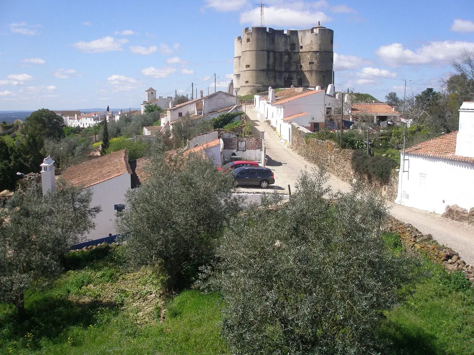 Whitewashed houses with red-tiled roofs line a lane climbing a hill among olive trees towards a massive grey stone keep with four round corner towers. Beyond the village, a plain stretches to the horizon under a blue sky.