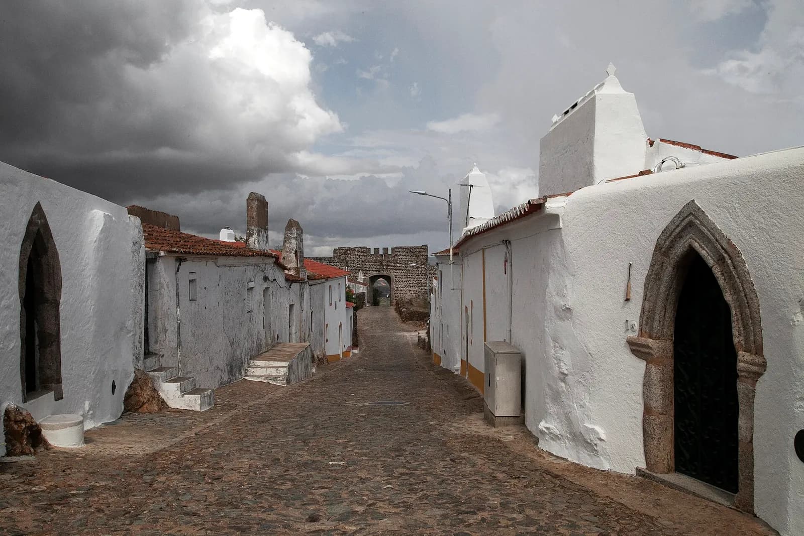 A cobbled street runs between low whitewashed houses towards a stone gateway with battlements under a heavy grey sky. On the right, a house has a pointed Gothic stone doorway.