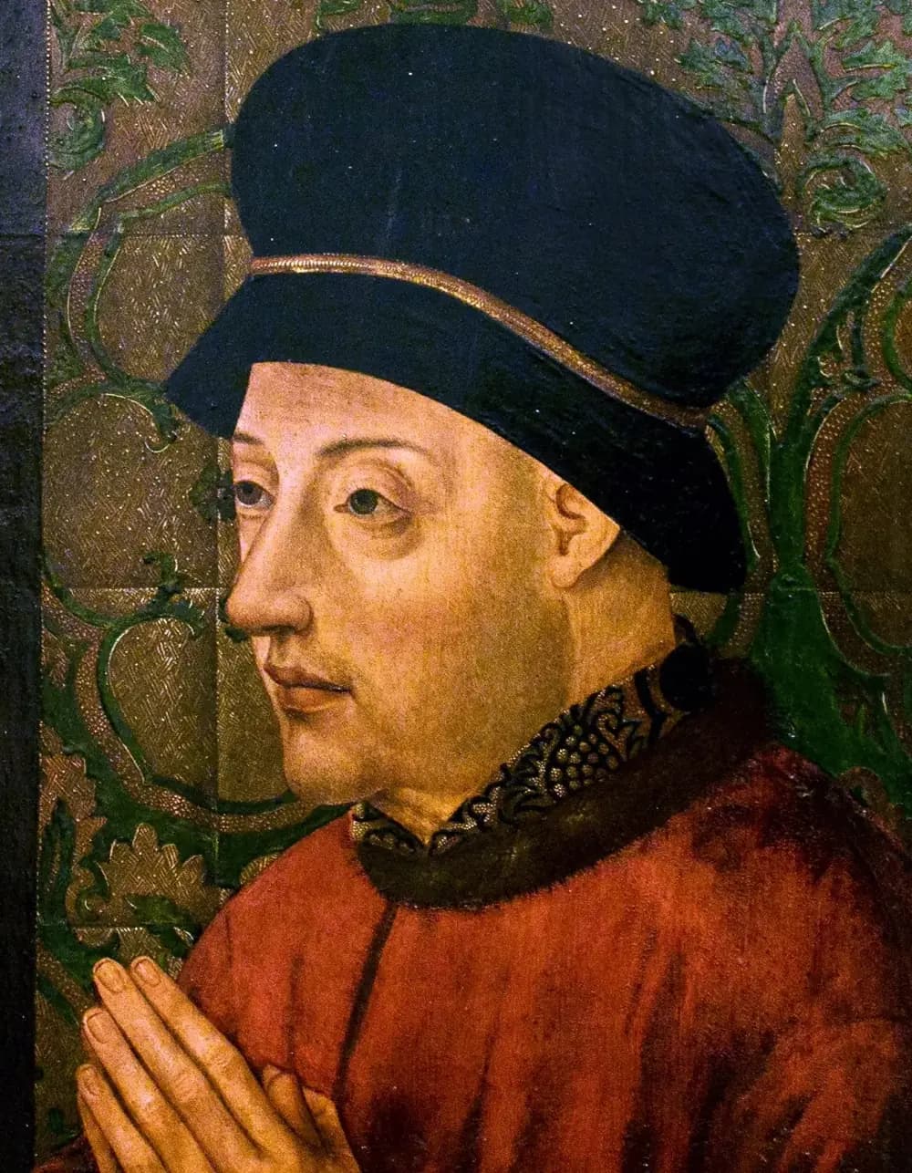 A painted portrait of a clean-shaven man turned to the left, with heavy-lidded eyes, wearing a tall dark blue hat with a gold band and a red robe with a fur collar, his hands joined in prayer against a green and gold patterned background.