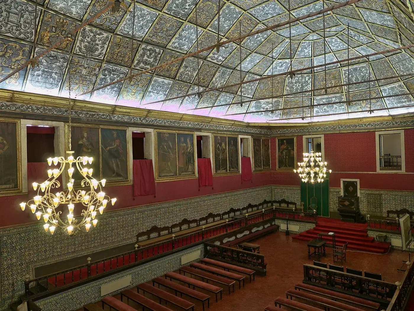 A tall hall seen from a gallery, with a painted coffered ceiling in grey and gold, red walls hung with full-length portraits of kings, blue and white tiles below, rows of benches and a raised dais with a chair at the far end, lit by two gilded chandeliers.
