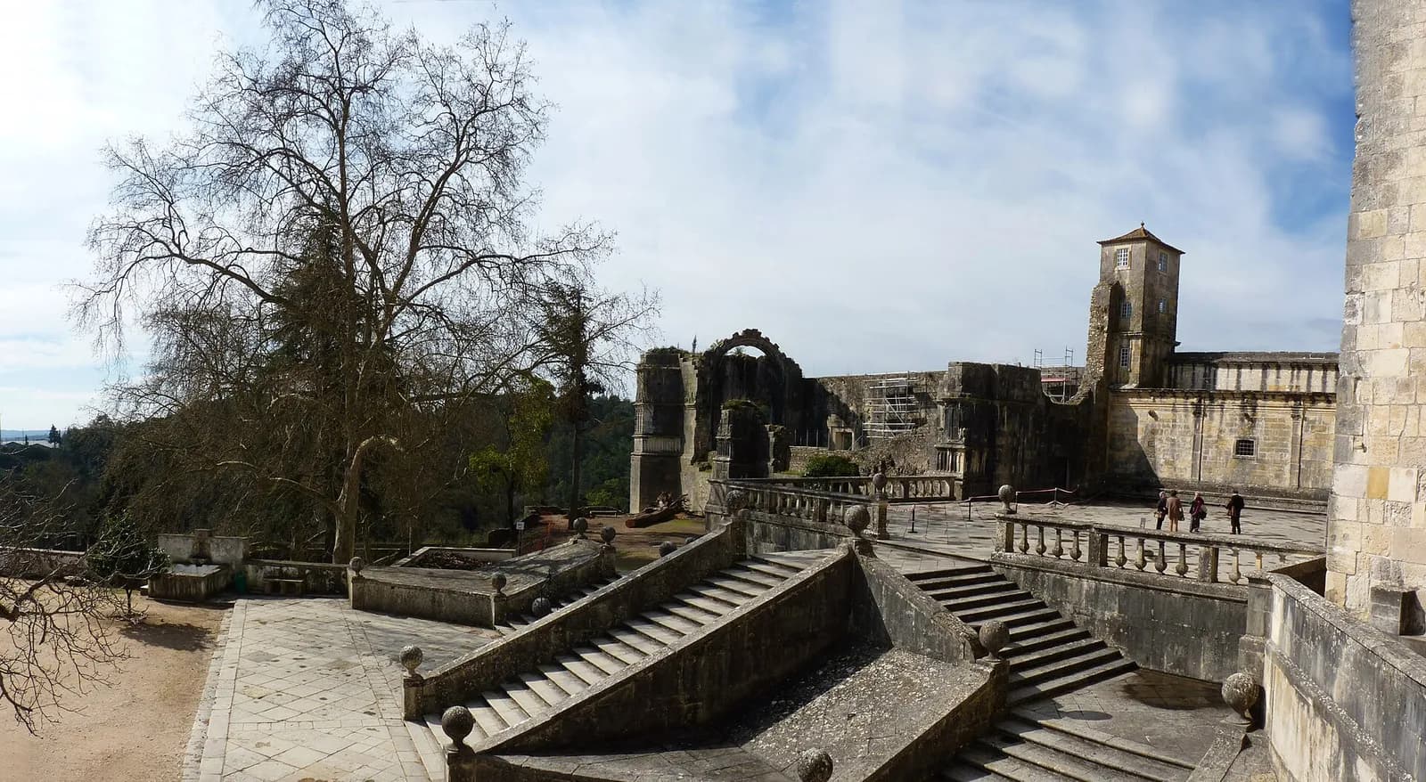 Stone terraces and balustraded double stairways at the Convent of Christ in Tomar under a pale blue sky, with a ruined arch, a square tower and a bare tree on the left.