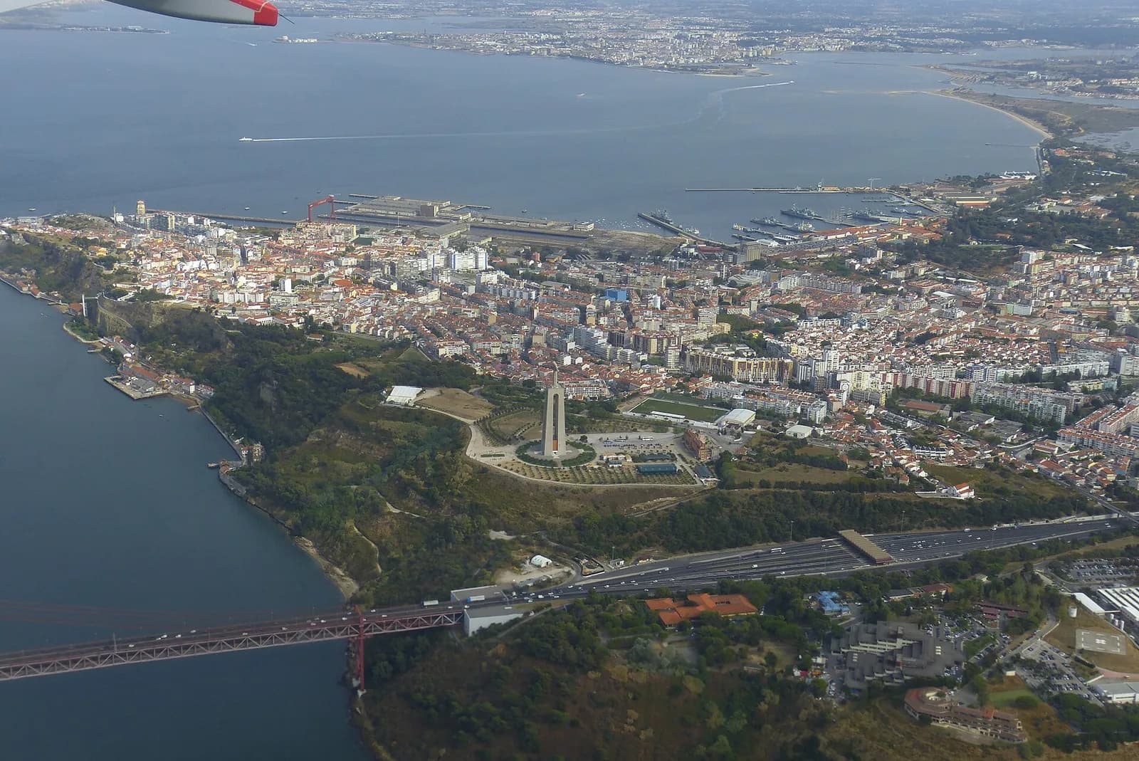 Aerial view of a wooded headland above a river. A pale tower stands at the centre of a circular plaza, with the dense streets of a town behind it, a shipyard and naval base on the waterfront beyond, a wide estuary at the top and a red suspension bridge crossing the water at the bottom left. The tip of an aircraft wing shows at the top left.