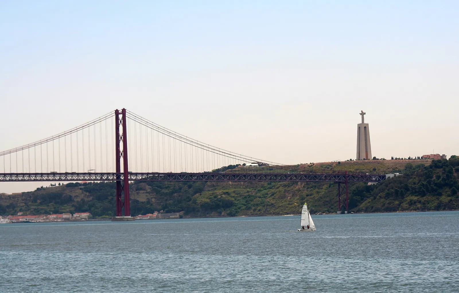 A wide river with a small white sailing dinghy in the foreground. On the left a red suspension bridge crosses to a green, wooded far bank. On the hilltop to the right stands a tall pale concrete tower on two legs, topped by a statue with outstretched arms.