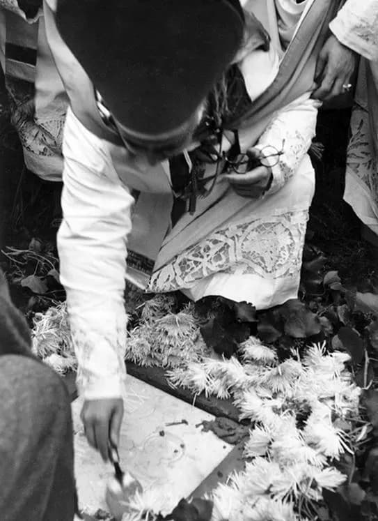 Black and white photograph, seen from above, of a cleric in a dark skullcap and a white lace surplice bending to spread mortar with a trowel on a stone slab edged with white chrysanthemums. Another cleric in lace vestments kneels beside him, holding a pair of glasses.