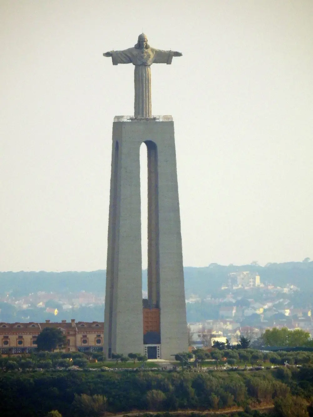 A tall pale concrete pedestal formed by two pillars joined at the top, with a narrow arched opening between them, carrying a statue of Christ with outstretched arms against a hazy sky. Wooded hills and buildings lie behind.