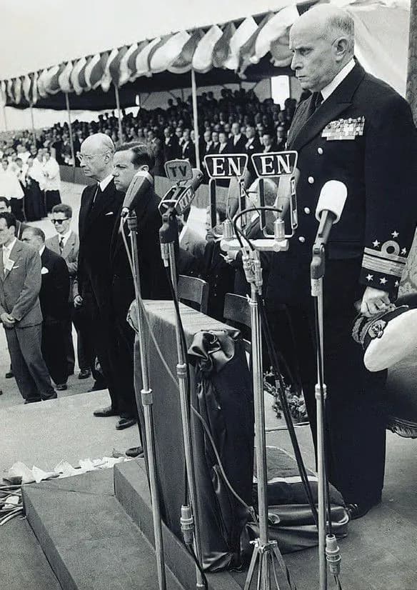 Black and white photograph of a bald man in an admiral's dress uniform with medals standing stiffly at a bank of microphones marked TV and EN. Men in dark suits stand beside him and a covered stand full of spectators fills the background.