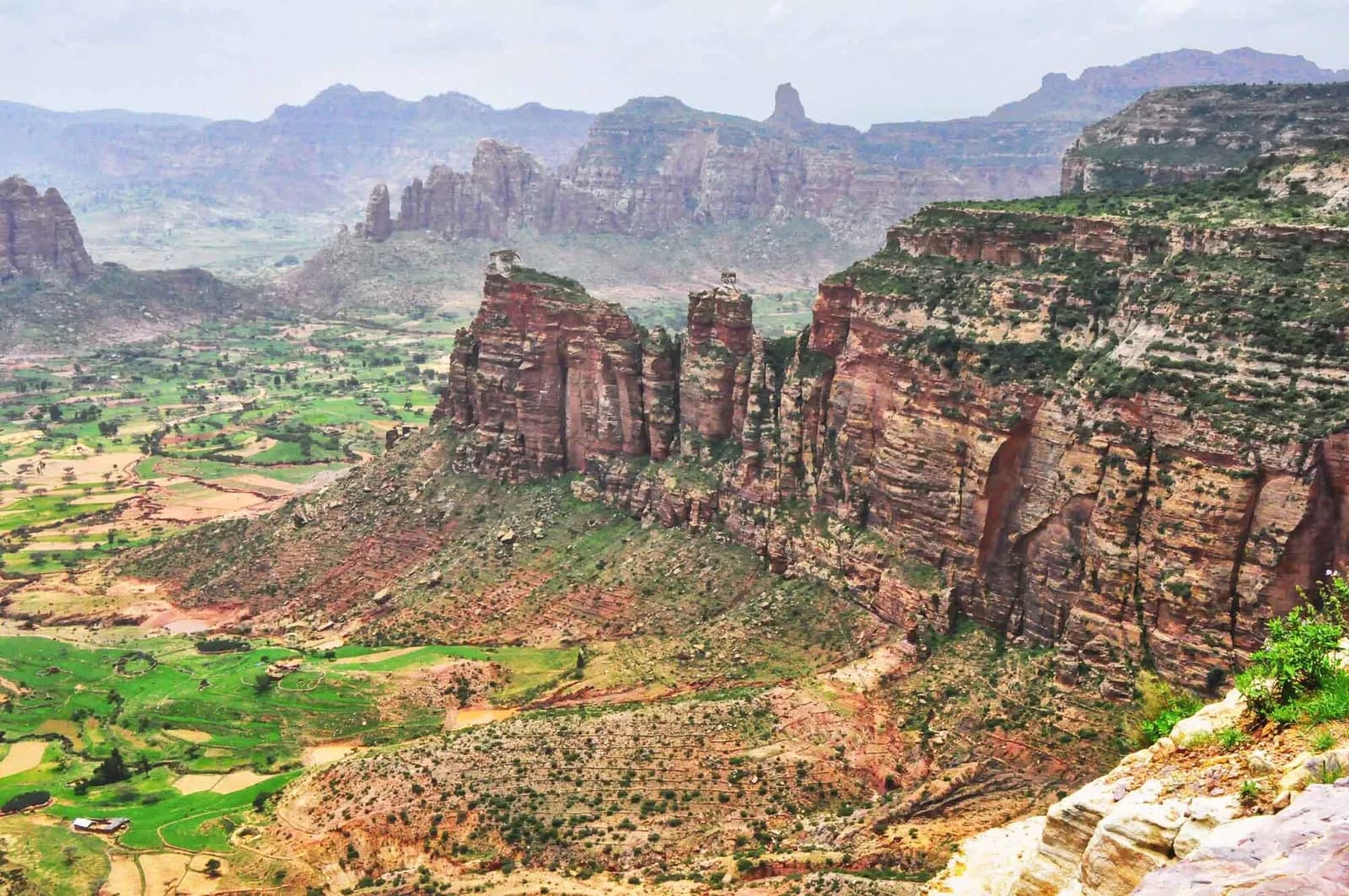 Red sandstone mountains with sheer cliffs and flat, scrub-covered tops rise above a valley of small green fields. Further ridges and pinnacles fade into haze behind them.