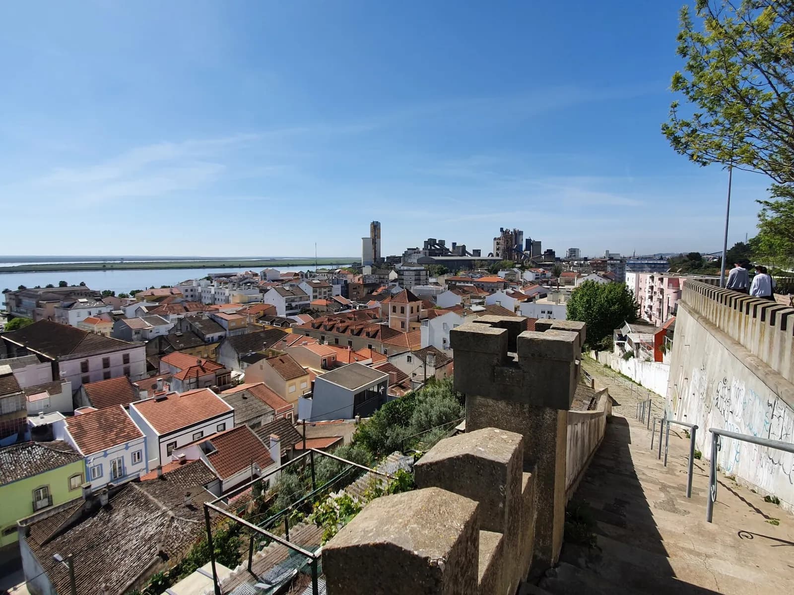 View over the tiled roofs of a small riverside town towards a wide, flat estuary under a blue sky, with the tall towers of a cement works on the right and a stone wall with battlements in the foreground.