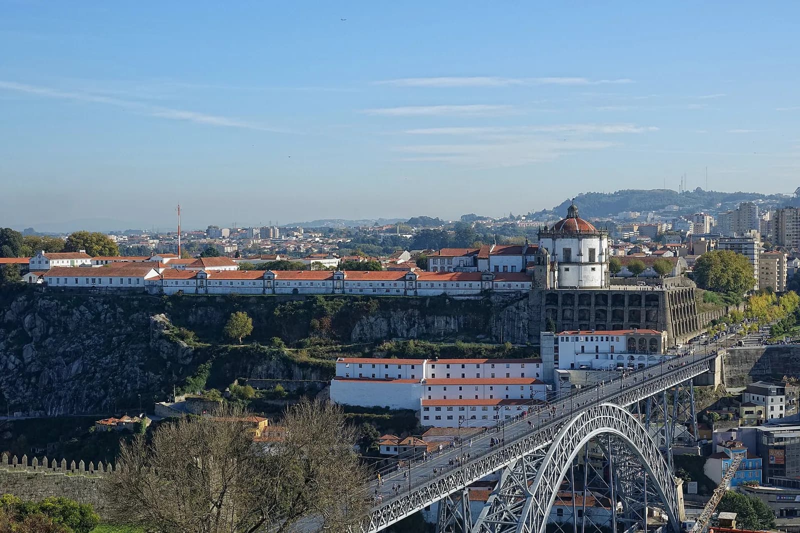 A long white building with a domed round church sits on top of a rocky cliff, with the upper deck of an iron arch bridge in the foreground and a town spreading behind.