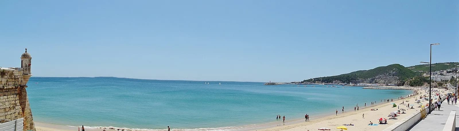 Panoramic view of a wide bay with calm turquoise water and a long sandy beach backed by a seafront promenade. A stone fortress wall with a round watchtower stands at the left edge.