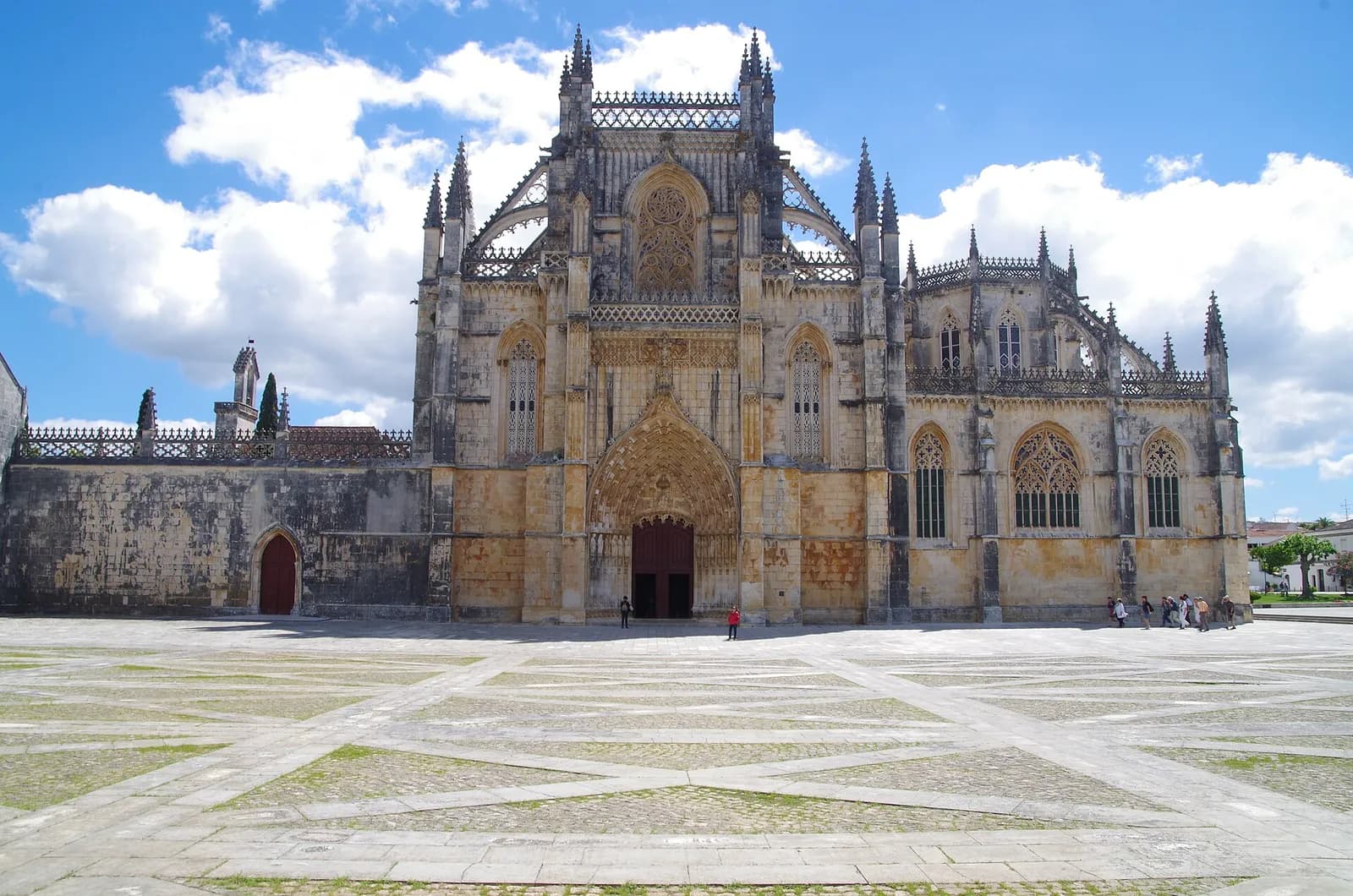 The west front of a large Gothic monastery church in pale golden stone, with pinnacles, tall traceried windows and an arched portal, seen across a wide paved square under a blue sky with white clouds.