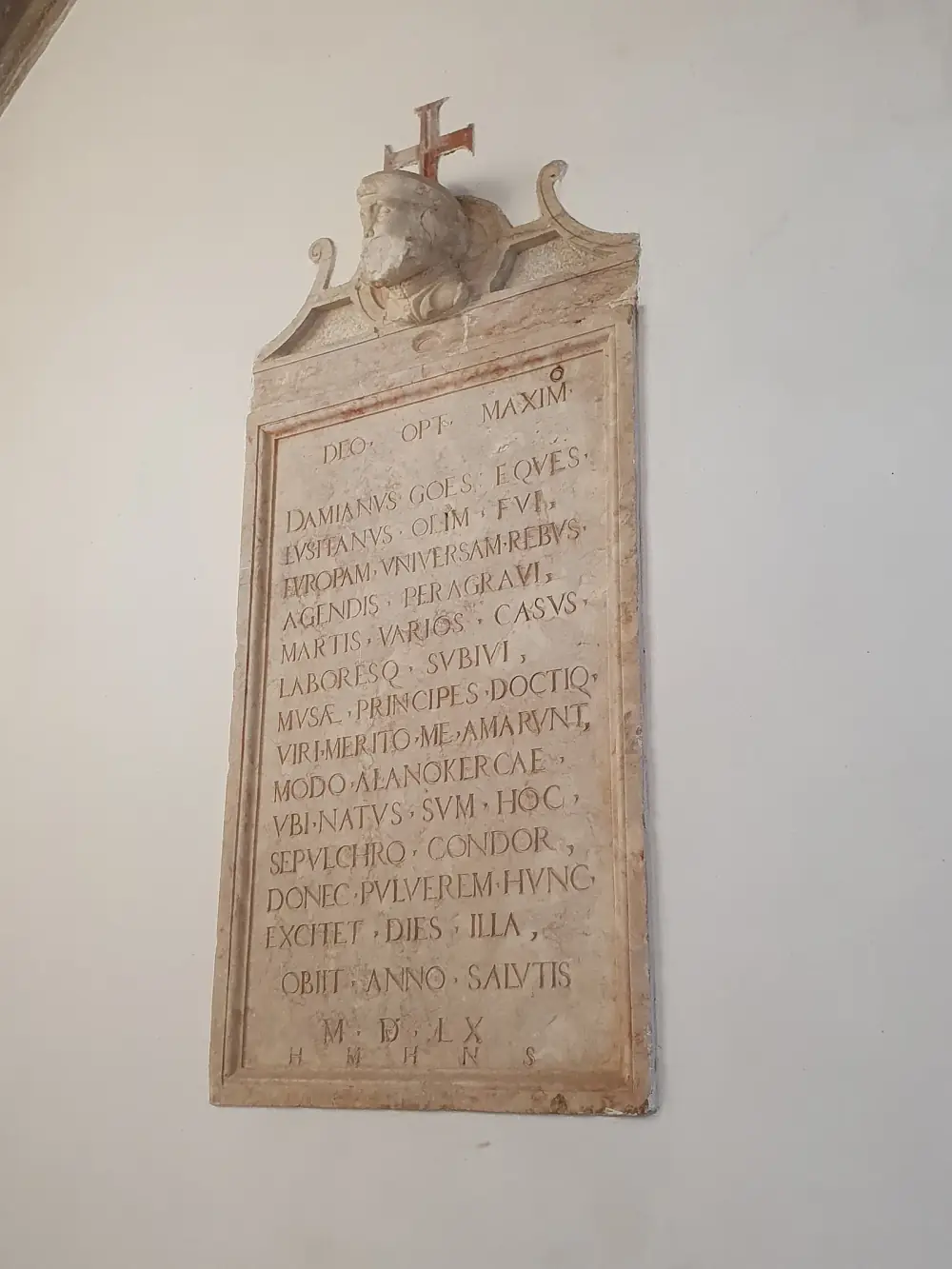 A tall stone tablet on a white wall, carved with a Latin inscription in capital letters and crowned by a small stone bust of a bearded man in a cap beneath a red cross.
