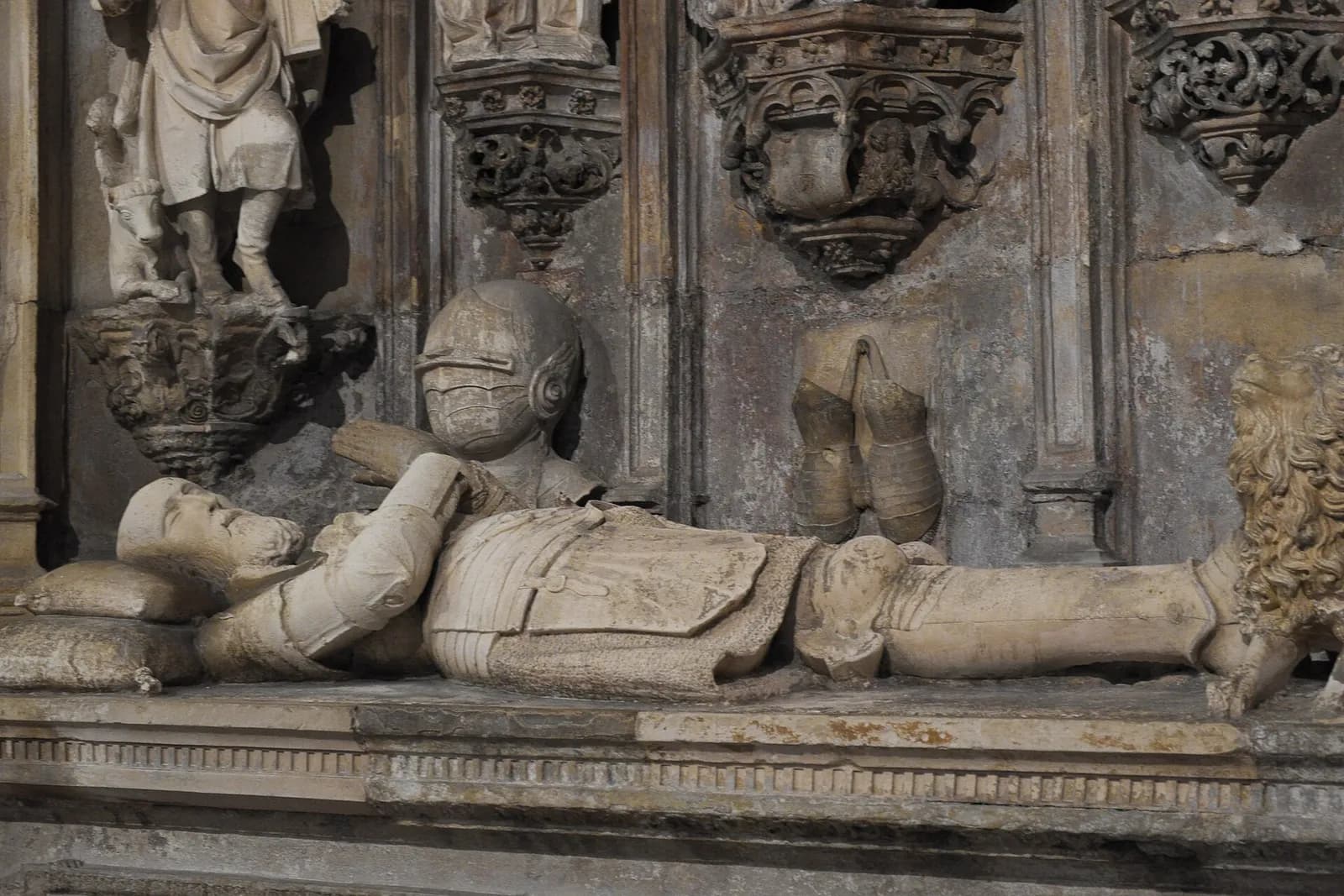 A pale stone figure of a king lies on a tomb chest, head on a cushion, hands raised in prayer, in armour with a crown. His helmet and gauntlets are carved on the wall behind him, below small statues and canopies, and a lion lies at his feet.