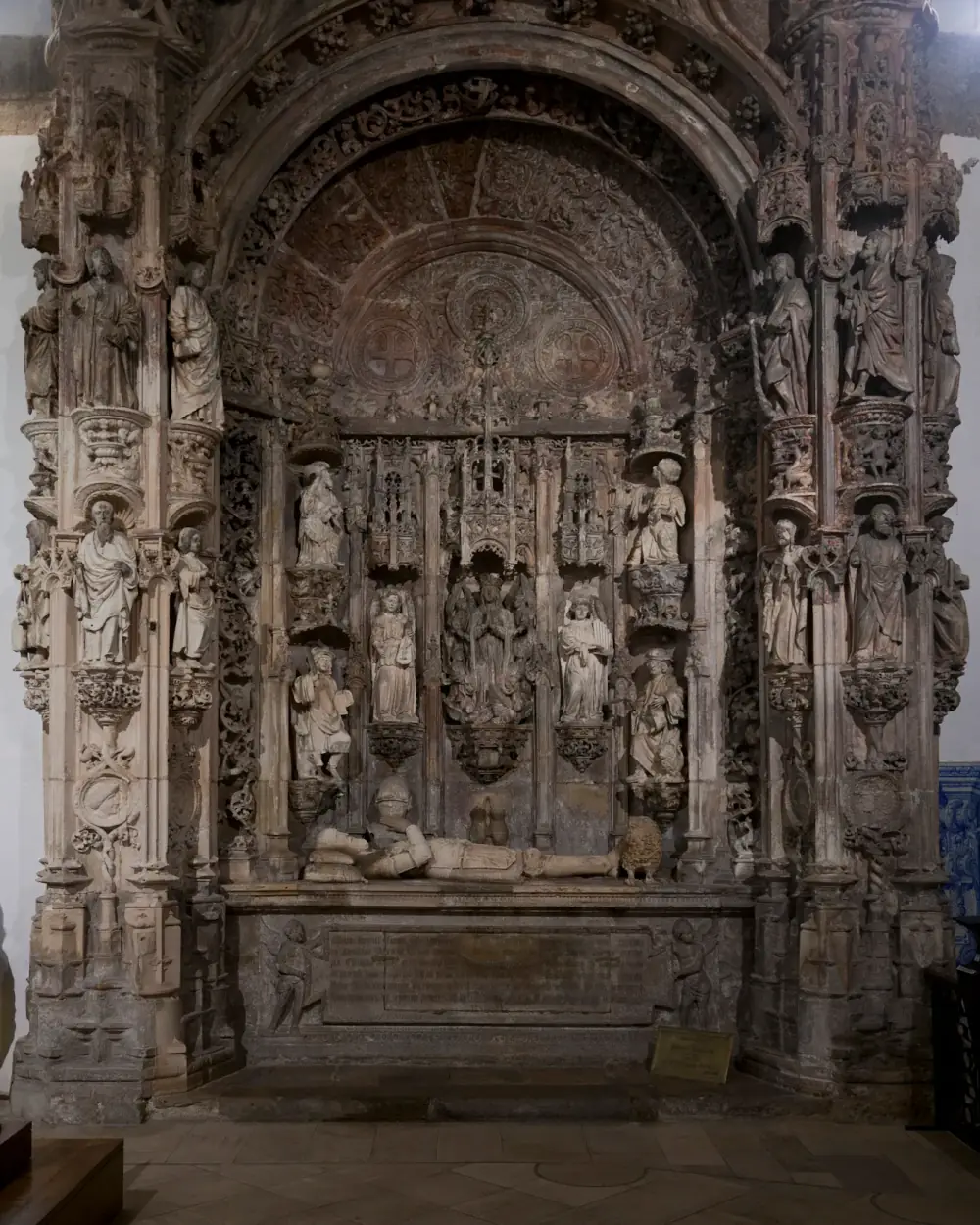 A towering carved stone tomb set into a church wall: a round arch framed by pinnacled piers covered in small statues in niches, with a reclining crowned figure on the tomb chest at the bottom and an inscription panel below it.